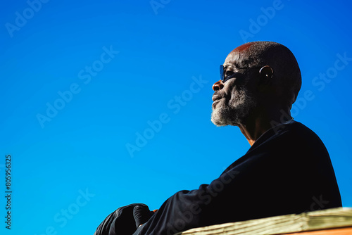 A focused middle-aged Afro-American man practicing yoga on a sun-drenched deck, his silhouette outlined against the backdrop of a clear blue sky.