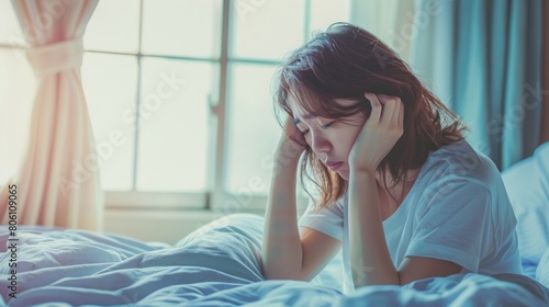 Tired asian woman sitting on bed with head in hands, feeling sick and unwell at home. Young girl suffering from falling ill or having handsome pain while alone during morning time in bedroom.