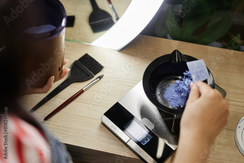 High angle closeup of hairstylist mixing bleach in plastic bowl and weighing solution on scales preparing to do hair coloring in beauty salon, copy space