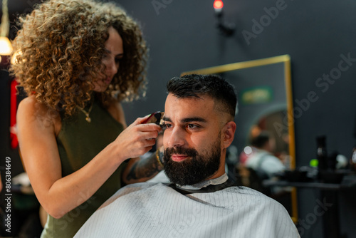 Female barber meticulously trims and styles a male client's hair with electric clippers in a modern barber shop, ensuring a precise and stylish cut.