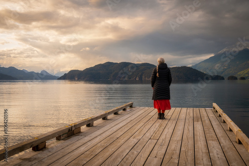 Senior woman gazing out over a lake at sunset from a boat dock. Seen at Harrison Lake, British Columbia, with dramatic clouds and calm waters.