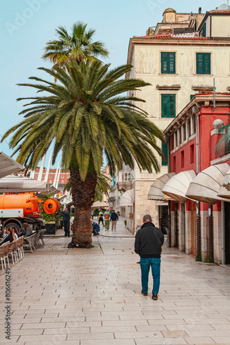 A man walking along the old town in Split near a palm tree | człowiek idący wzdłuż starego miast w Splicie nieopodal palmy | čovjek šeće starom gradskom jezgrom u Splitu u blizini palme