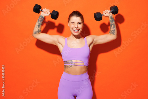 Young fitness trainer instructor woman sportsman wear purple top clothes spend time in home gym hold dumbbells raise up hands wink isolated on plain orange background. Workout sport fit abs concept.