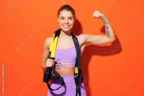 Young fitness trainer woman wears top shorts purple clothes in home gym hold trx loops do Total Body Resistance Exercise show biceps isolated on plain orange background. Workout sport fit abs concept.