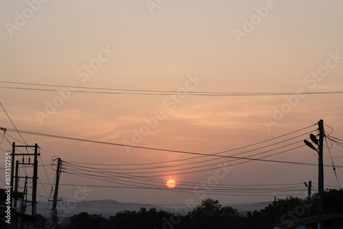 Low setting full round sun at sunset framed by electricity and telephone cable wires and trees, Accra, Ghana