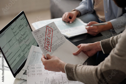 Hands of young unrecognizable male owner of apartment taking financial bill out of envelope while sitting by table with laptop and papers