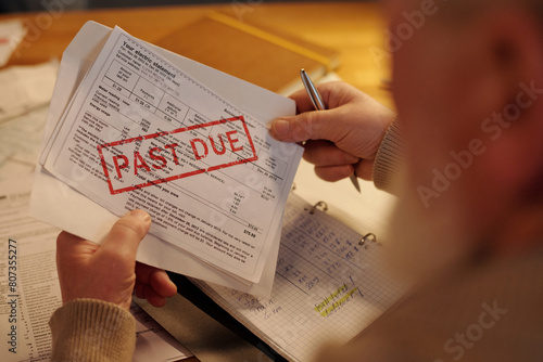 Hands of aged man holding silver pen, envelope and unpaid financial bill with past due stamp while sitting by table and checking post