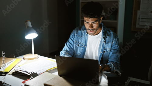 Handsome hispanic man working late on laptop in office, concentrated under desk lamp light.