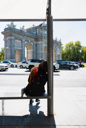 The Alcala Door (Puerta de Alcala) is a one of the ancient gate of the city of Madrid - A young girl waiting for a bus at a bus stop - Madrid, Spain