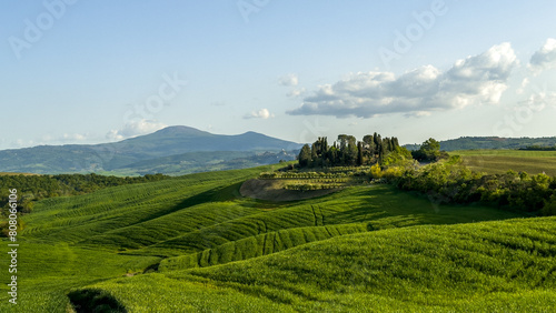 zachód słońca wydłuża cienie padające ze wzgórza na którym wybudowano piękna willę. w dole rozległe pola uprawne. Toskania, Włochy