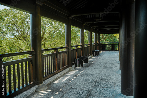 The image shows a long, covered walkway with wooden benches and a view of a lush green garden.