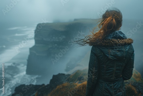 Alone in contemplation, a person stands on a grassy outcrop, gazing at the mist-enveloped sea cliffs and waves