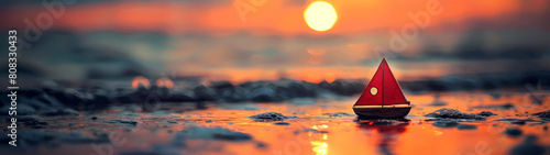 At the water's edge, a diminutive toy sailboat lies peacefully upon the dampened grains of beach sand, its miniature form basking in the radiant embrace of the sunset's warm orange light