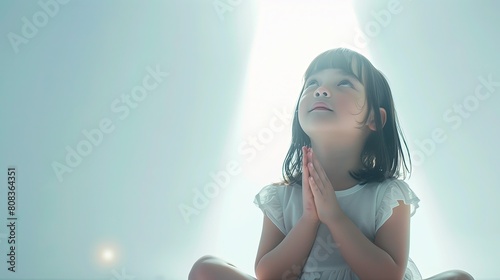Mała dziewczynka, siedząca na podłodze na jasnym tle, modli się z zamkniętymi oczami i złożonymi dłońmi