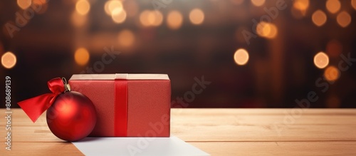 Red Christmas gift box with ribbon red ball hanging and a blank notepaper on the table providing an ideal copy space image for your text
