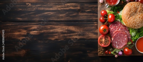 A top down copy space image of a breakfast consisting of a burger with salami and vegetables accompanied by tomato juice placed on a wooden background