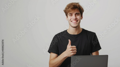 Smiling young slim businessman entrepreneur male wearing plain black t-shirt looking from laptop while giving thumbs up hands isolated on white background
