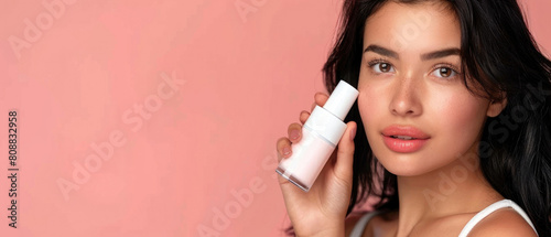 young beautiful girl with black hair and perfect smooth skin posing in front of the camera and holding a white cosmetic bottle mockup in pastel colored studio