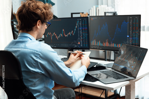 Working young business trader looking on laptop on screens, holding notepad and pen on market stock graph investing in real time at office on panorama, analyzing dynamic investment concept. Gusher.