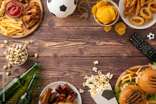 Football fan's delight. Top view of snack assortment: crackers, onion rings, popcorn, wings, hamburgers, nuts, beer, soccer ball, referee gear, remote control, on wooden desk with empty space for text