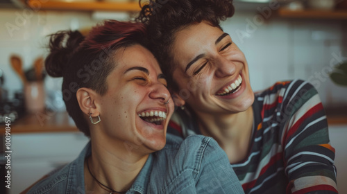 Young queer couple laughing together indoors. Happy young queer couple having fun together while standing in their kitchen. Romantic young LGBTQ+ couple bonding fondly at home. Stock Photo photography