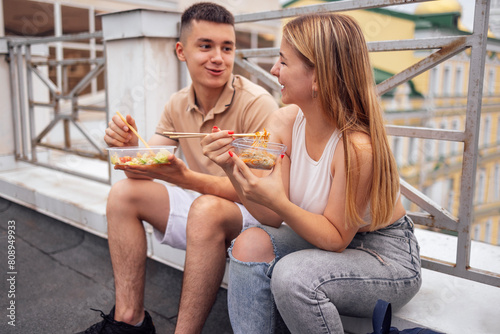 Two teenagers are sitting on the parapet on the roof of the house and eating Asian food