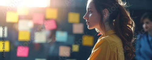 Young woman in yellow jacket contemplating project ideas on a sticky note wall. Strategic planning and creativity concept