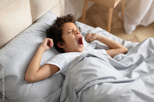 African American young boy is laying in bed, his mouth wide open in a yawn. He appears tired as he stretches his arms out. The background shows a cozy bedroom setting.