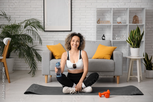 Happy mature sporty woman with dumbbells and bottle of water sitting at home