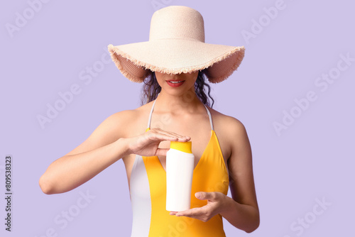 Beautiful young African-American woman in swimsuit and wicker hat with bottle of sunscreen cream on lilac background