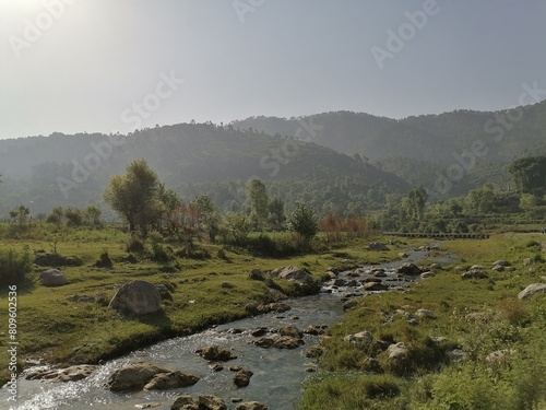 A gentle brook babbles and winds its way through the heart of the lush green mountains, its crystal clear waters reflecting the vibrant hues of the surrounding landscape