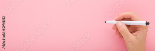 Young adult woman hand holding black white color pen and writing on light pink table background. Pastel color. Closeup. Wide banner. Empty place for text. Top down view.