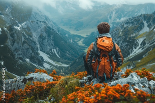 Solo traveler with backpack stands contemplating the vast mountain landscape with orange flowers in foreground