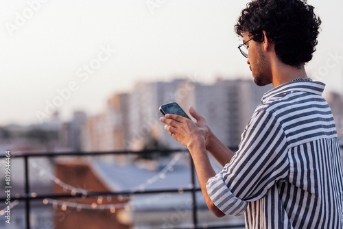 Young curly-haired guy in glasses and striped shirt answers video call on phone. Smiling mixed-race man shows victory symbol and takes selfie standing on roof of house. Big city on background.