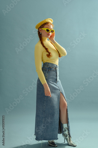 Fashionable redhead woman wearing yellow beret, sunglasses, turtleneck, maxi denim skirt, silver ankle boots, posing on blue background. Studio full-length fashion portrait