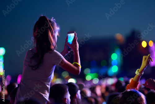Girl films music concert on smartphone amid lively festival crowd at twilight. Audience enjoys live performance, memories, vibrant stage lights beam. Fans wristbands share experience social media.
