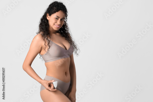 Young African-American woman in underwear with menstrual cup on light background