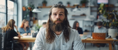 An attractive long-haired bearded manager with his laptop computer sits at his desk in a creative office. His colleagues work in the background.