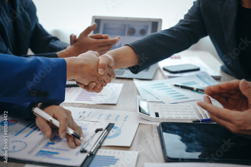 In a business team meeting, success is celebrated with a handshake. Hands close up, dressed in formal suits, working at desks with financial papers, calculators, and laptops.