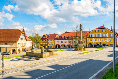 A picturesque view of a peaceful square in Zacler, Czech Republic. Colorful buildings with classic European architecture line the street. A beautiful fountain with an intricate statue is at the center