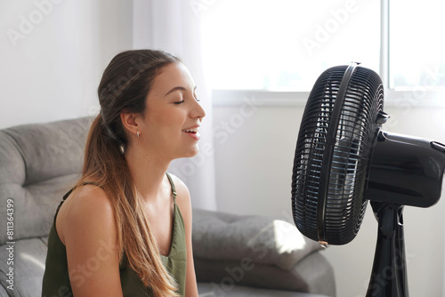 Happy smiling young woman sitting on couch in sunny hot summer day enjoying fresh air in the front of electric working fan