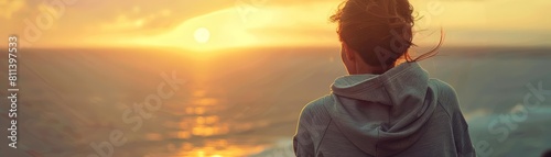 A sporty woman in a sweatshirt gazes out at the ocean, embracing the warmth of the setting sun