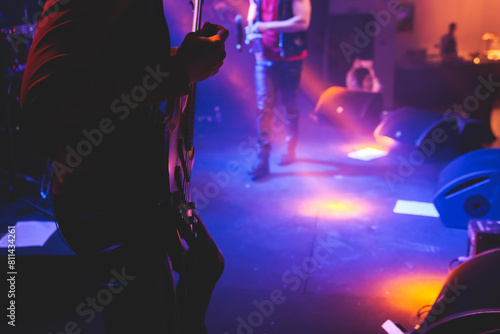 Concert view of an electric guitar player with vocalist and rock band performing in a club, male musician guitarist on stage with audience in a crowded concert hall arena, hands on a bass guitar