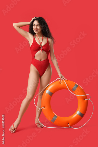 Beautiful young happy African-American female lifeguard with ring buoy on red background