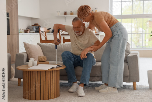 Mature woman helping her husband with crutch to stand up from sofa at home