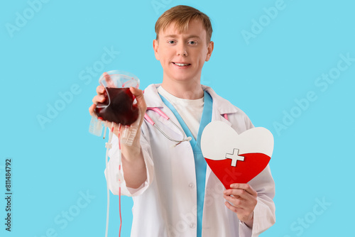 Male doctor with blood pack for transfusion and paper heart on blue background