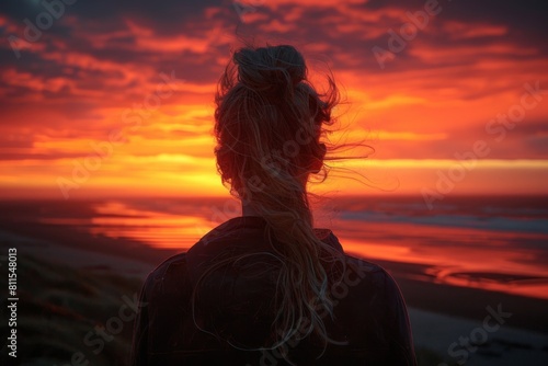 A woman facing a stunning sunset at the beach, her hair flowing in the breeze, evoking peace and reflection.
