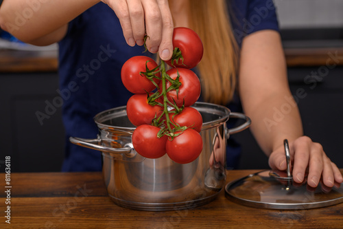 Kucharka trzyma gałązkę czerwonych dojrzałych pomidorów nad stalowym garnkiem do gotowania, zupa pomidorowa 
