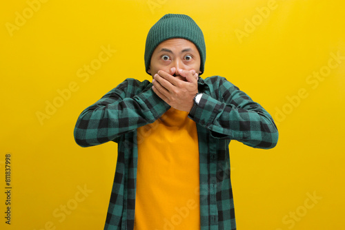 A surprised young Asian man, dressed in a casual shirt and sporting a beanie hat, is signaling for quiet by covering his mouth with his hands and shushing, while standing against yellow background.