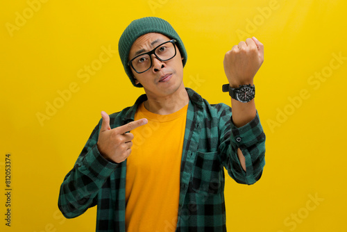 Frowning Asian man in a beanie and casual shirt impatiently taps his wristwatch, glancing at the camera with frustration. Clearly stressed by a deadline delay. Isolated on a yellow background